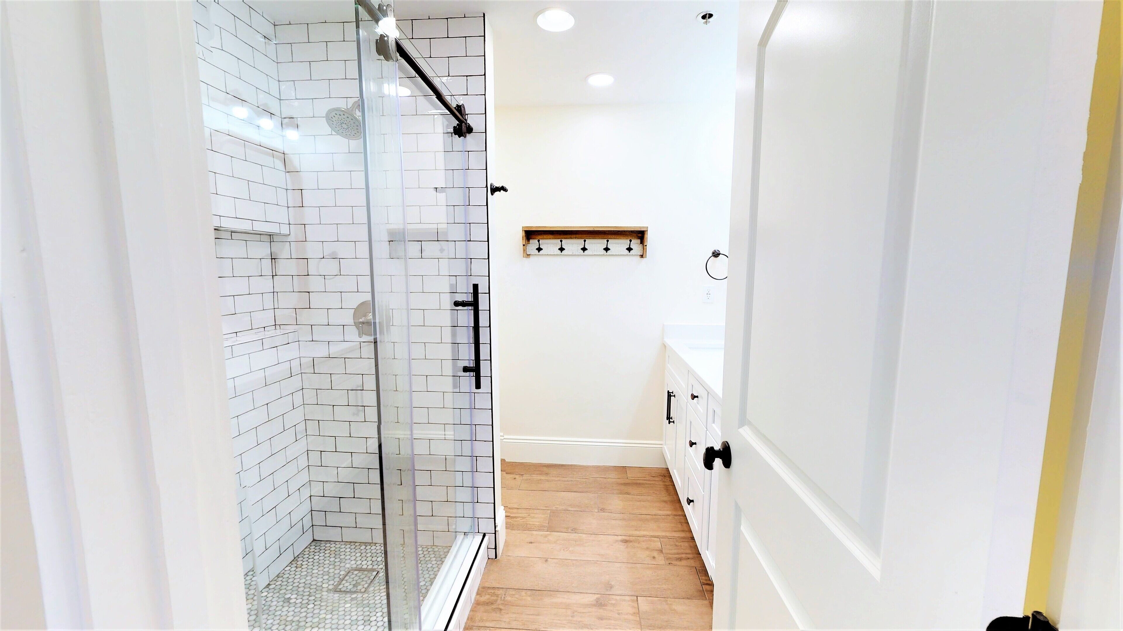 Un bagno moderno con cabina doccia in vetro, pareti rivestite di piastrelle bianche, pavimento in legno e un mobile lavabo bianco con cassetti.
