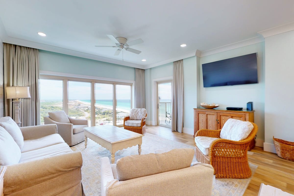 A spacious living room with a large window offering a view of the beach, a ceiling fan, and a flat-screen TV mounted on the wall.