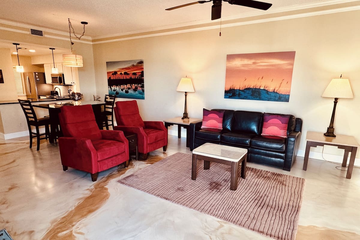 A living room with a black leather sofa, red chairs, a coffee table, and a rug. There is a painting on the wall and a ceiling fan.