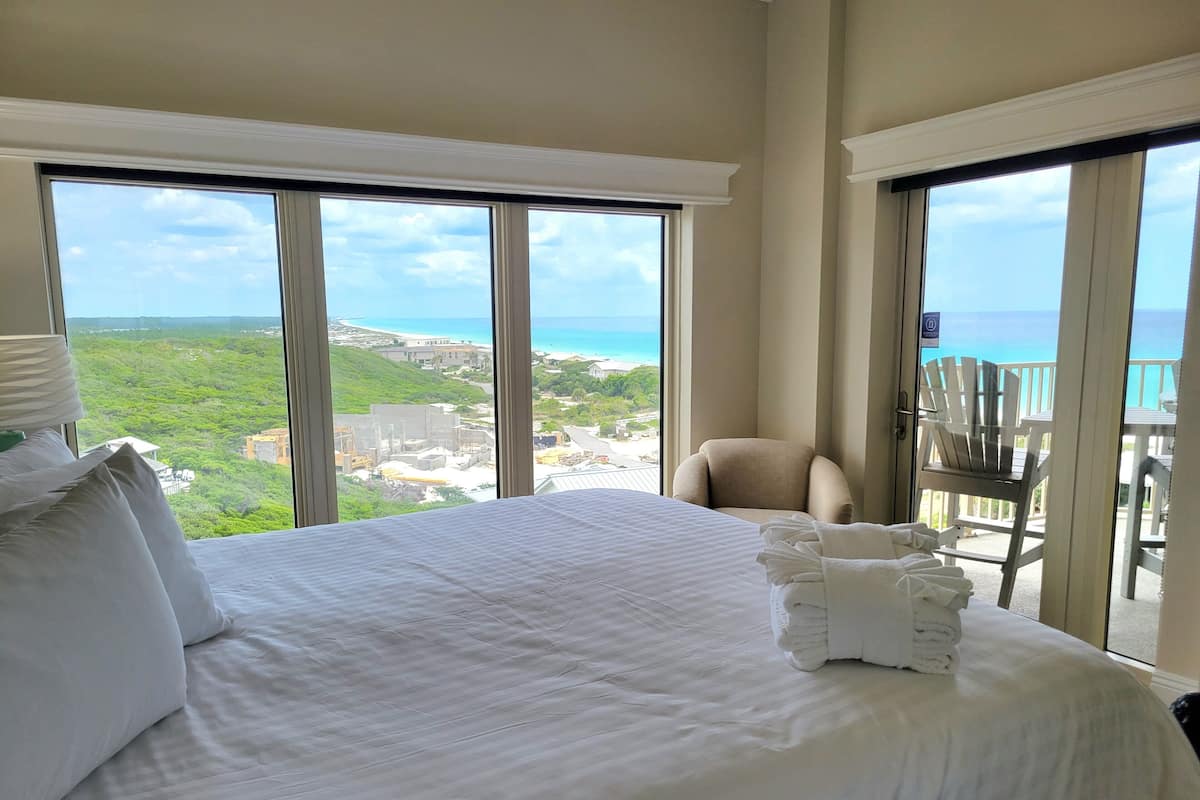 A hotel room with a large bed, a chair, and a balcony with a view of the ocean and greenery.
