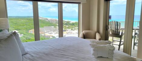 Habitación de hotel con una cama grande, una silla y un balcón con vistas al océano y al follaje.