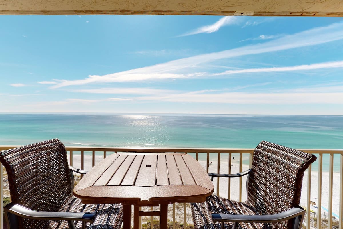 A wooden table and two wicker chairs on a balcony with a view of the ocean.