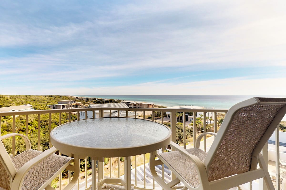 A balcony with a round table and two chairs, overlooking a beach and ocean.