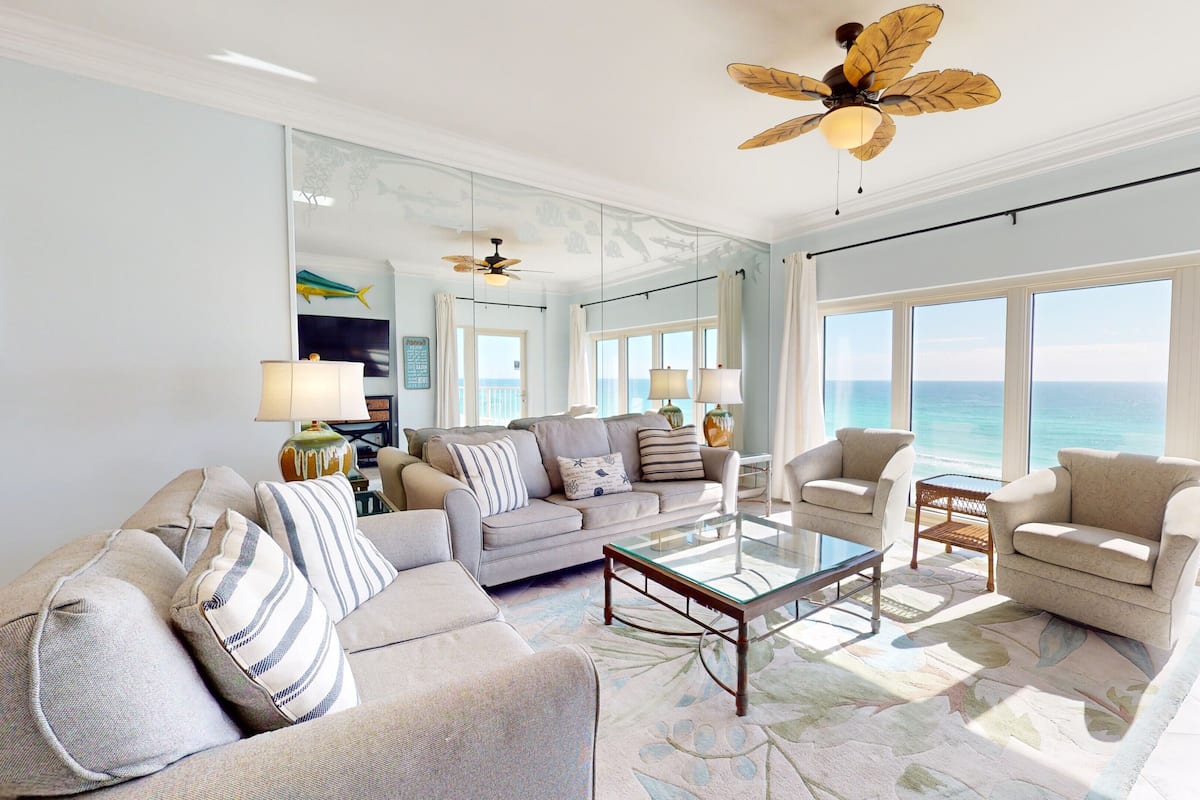 A spacious living room with a large window overlooking the ocean, a ceiling fan, and a modern coffee table.