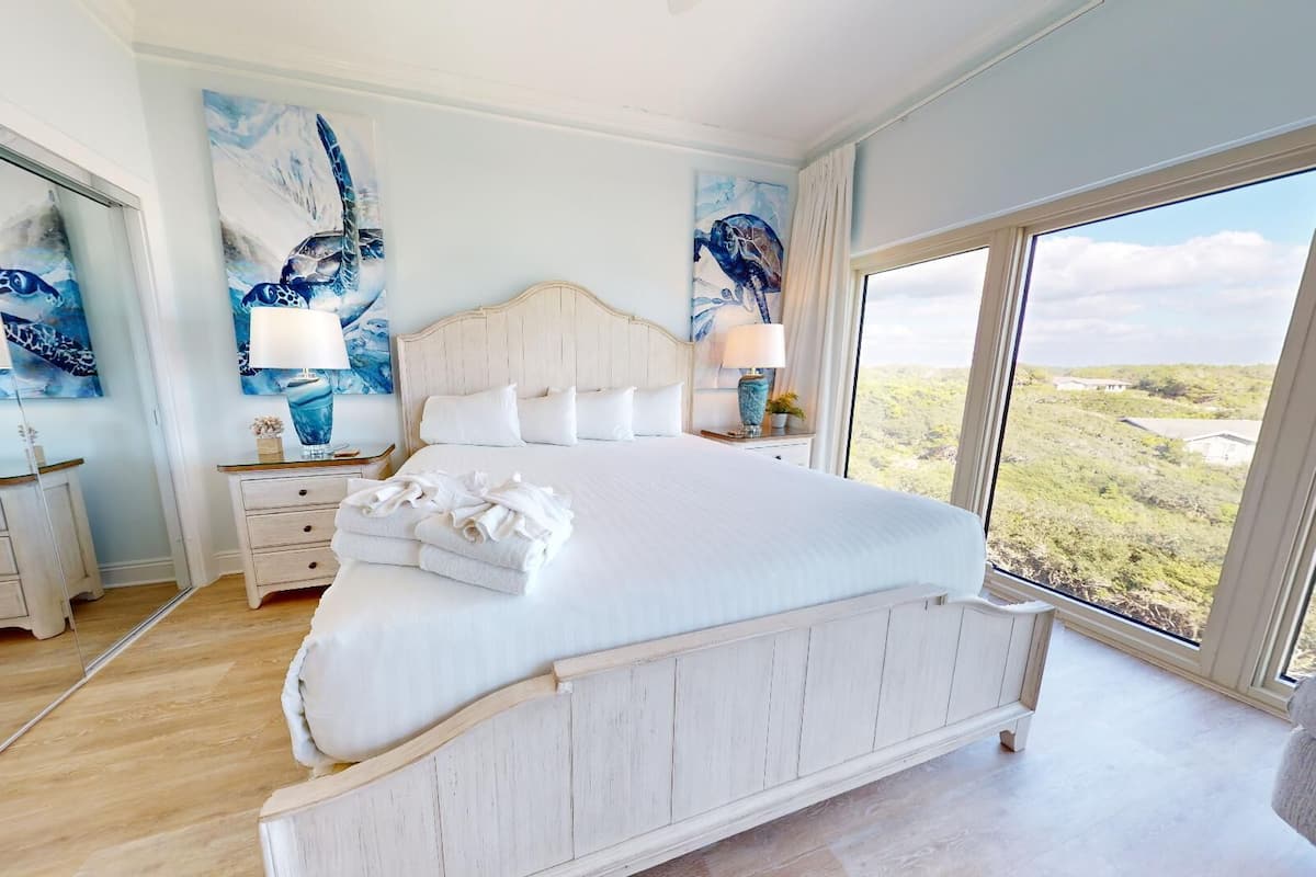 A spacious bedroom with a large bed, two bedside tables with lamps, and a view of the landscape through large windows.