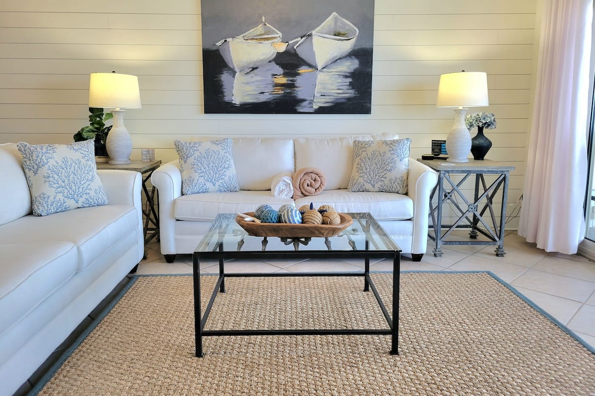 A living room with a white sofa, two side tables with lamps, a coffee table, and a painting of boats on the wall.