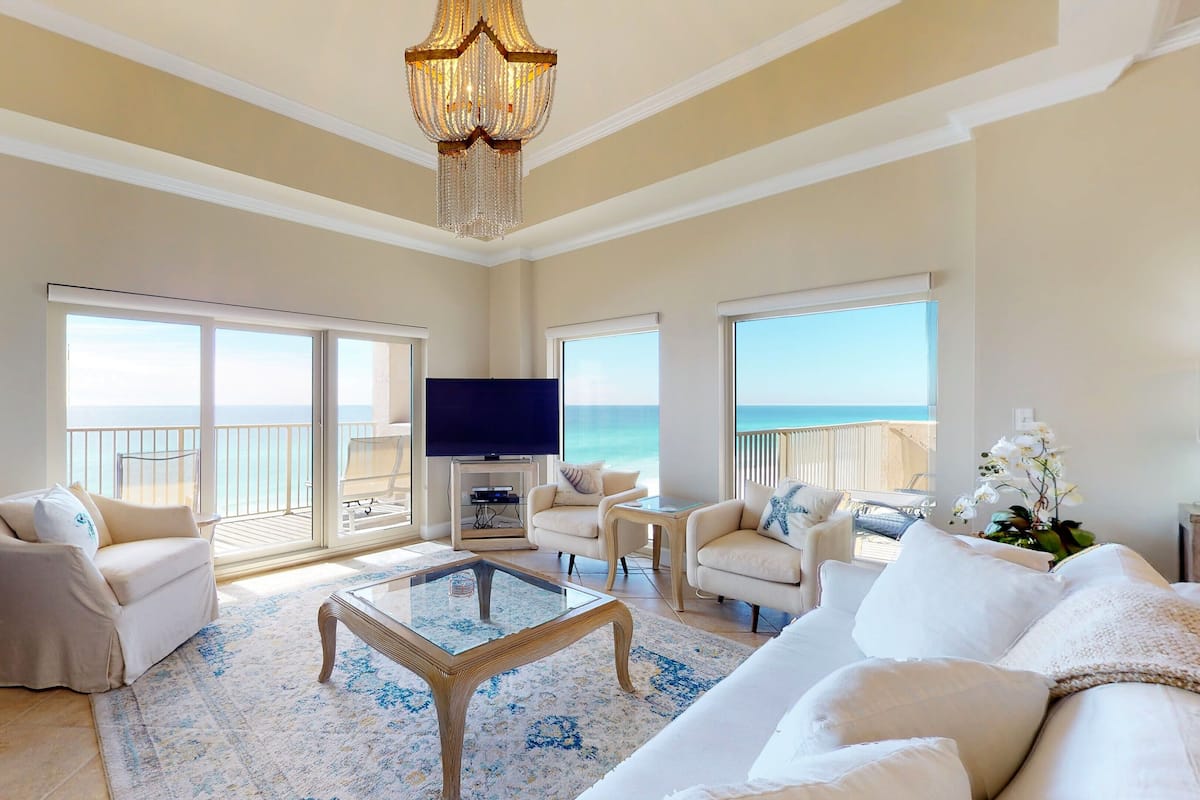 A spacious living room with a large TV, comfortable seating, a glass coffee table, and a balcony with a view of the ocean.