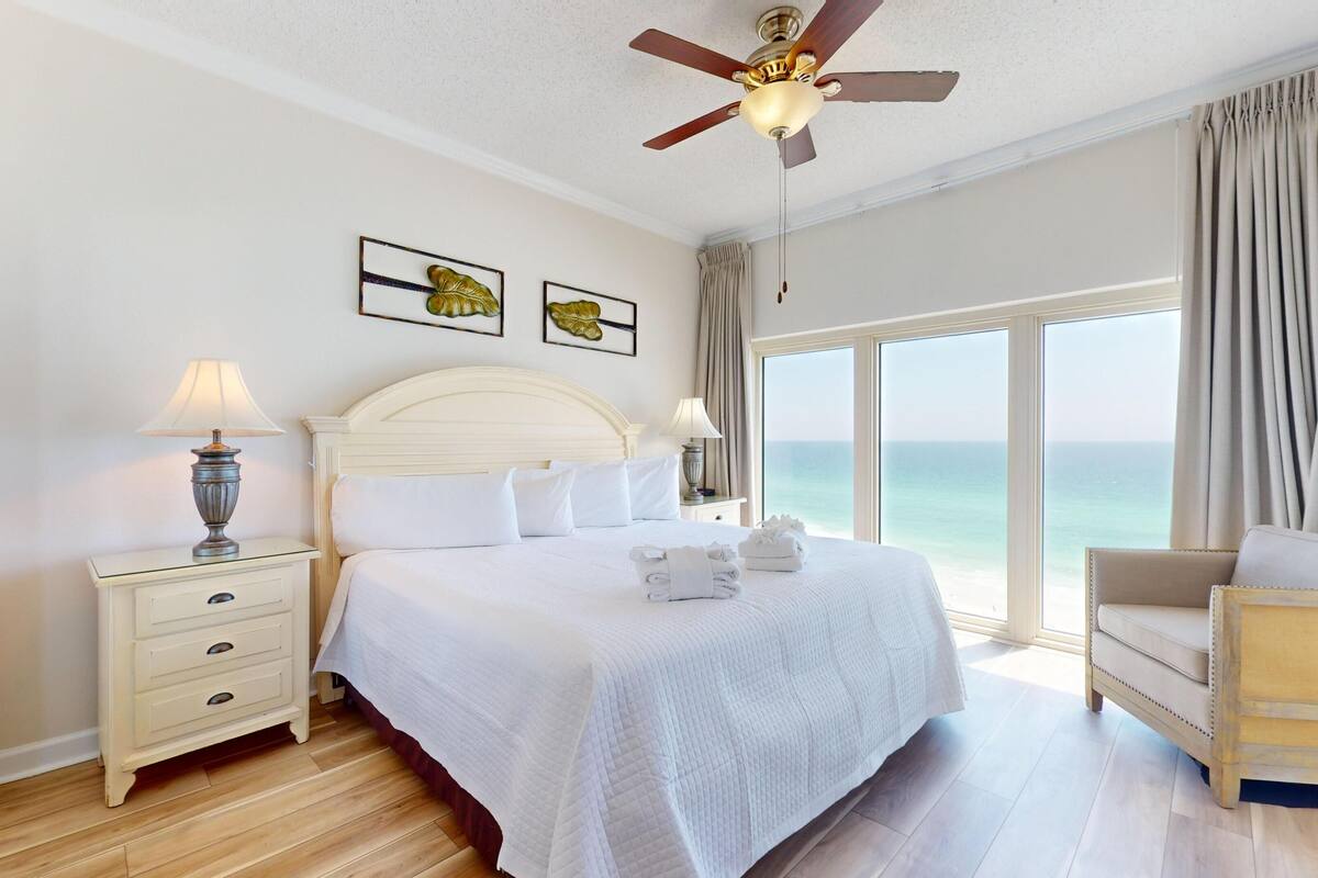 A hotel room with a bed, two bedside tables with lamps, a ceiling fan, a view of the ocean, and a sofa.
