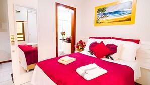 Hotellihuone, jossa on sänky, yöpöytä, peili, rantamaisemaa esittävä taulu ja ovi.