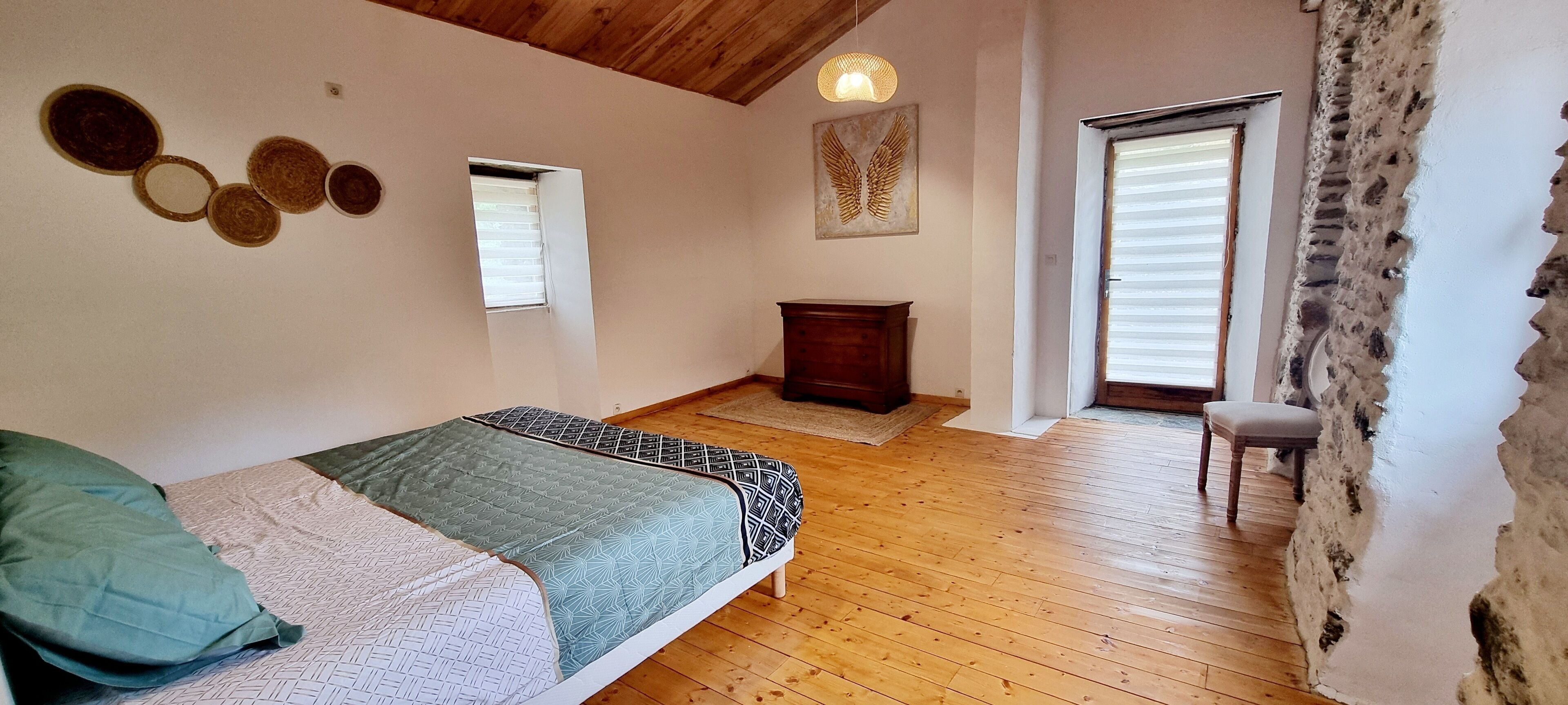 A bedroom with a wooden floor, a bed with patterned bedding, a small wooden chair, and a window with blinds.