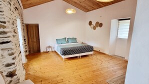 A bedroom with a wooden floor, a bed with patterned bedding, a small stool, and a wall-mounted light fixture. - Mas de 130m2 en Plein Cœur des Cévennes sur 12 Hectares (Lamelouze)