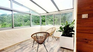 A room with large windows, wicker chairs, and a potted plant. - Mas de 130m2 en Plein Cœur des Cévennes sur 12 Hectares (Lamelouze)
