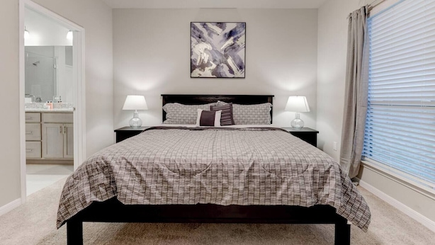 A bedroom with a bed, two bedside lamps, a nightstand, a mirror, and a window with blinds.