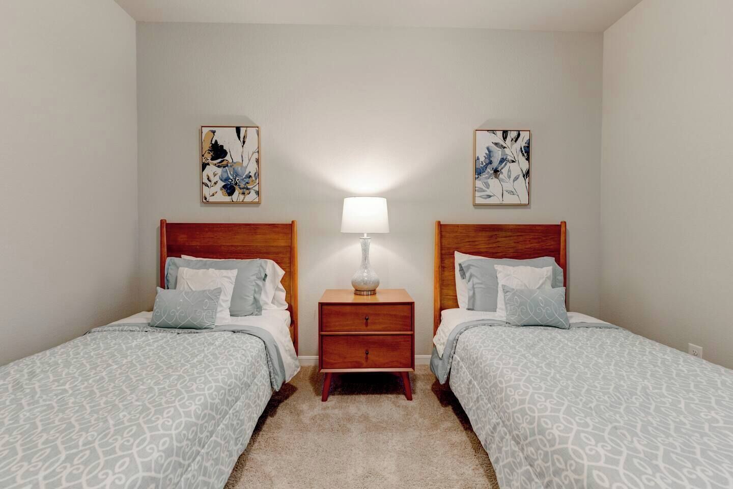 Una habitación con dos camas individuales, una mesita de noche con lámpara y dos cuadros colgados en la pared.