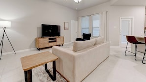 A modern living room with a sofa, a TV on a wooden stand, a ceiling fan, and a dining area with chairs.