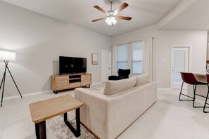 A modern living room with a sofa, a TV on a wooden stand, a ceiling fan, and a dining area with chairs.