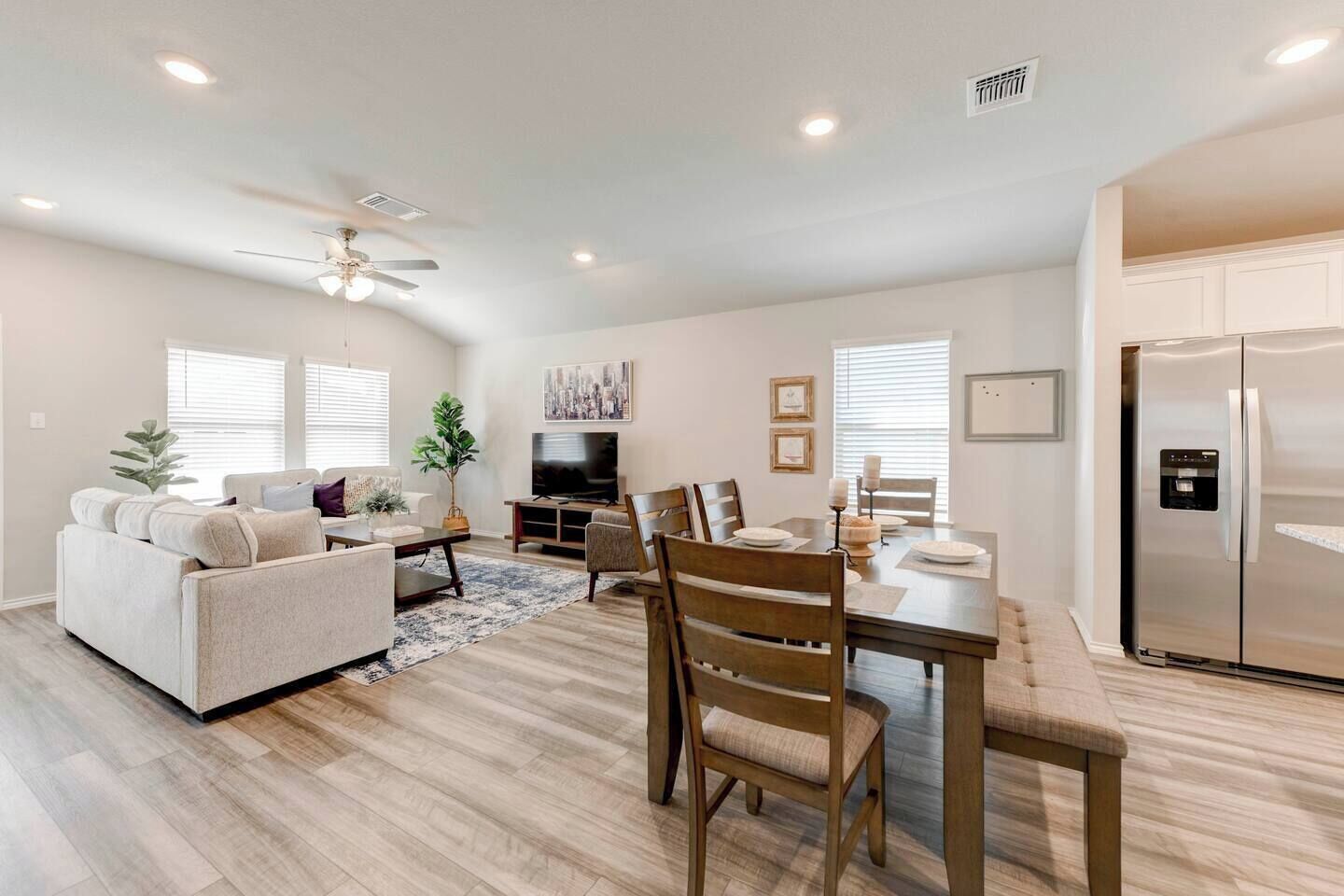A modern living room with a sofa, coffee table, and TV, a dining area with chairs and a table, and a kitchen with a refrigerator.