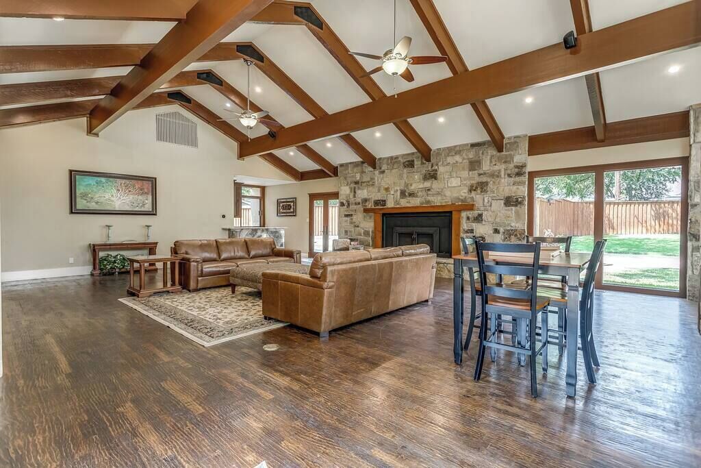 A spacious living room with a stone fireplace, wooden beams, and a large window overlooking a backyard.