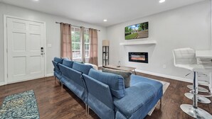 A living room with a sofa, a TV mounted on the wall, a fireplace, and a dining area with a bar. - Charming 3-bedroom bungalow with WiFi in Bham, 12 mins from UAB. (Birmingham)