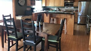 A kitchen with wooden cabinets, a stainless steel refrigerator, a dining table with chairs, and pendant lights.