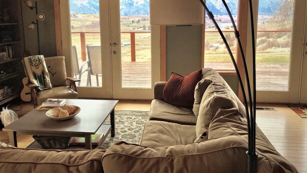 A cozy living room with a sofa, a coffee table, and a bookshelf. There are large windows with a view of a deck and mountains.