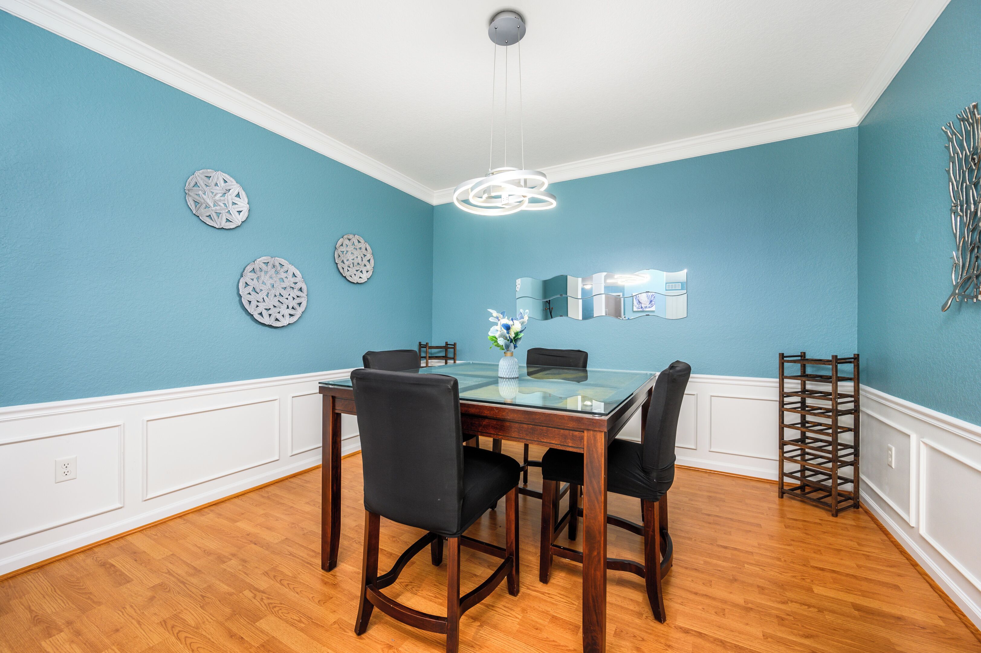 A dining room with a glass-top table, four chairs, decorative wall art, and a modern pendant light.