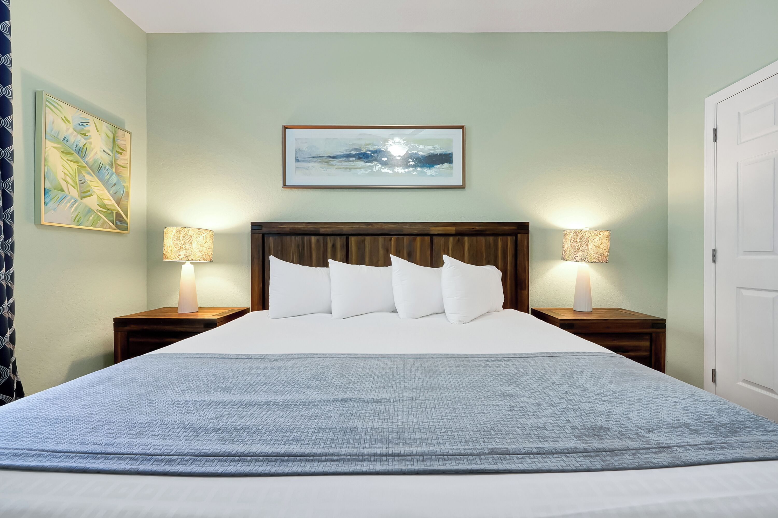 Una cama bien hecha con sábanas blancas y una manta azul con textura, flanqueada por dos mesitas de noche con lámparas, un cuadro abstracto enmarcado sobre la cama y una puerta a la derecha.