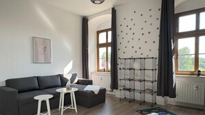 A modern living room with a grey sofa, a small white table, and a floor lamp. There is a window with curtains, a radiator, and a wall-mounted shelf with a lamp. - Ferienwohnung Plohn_immo-franzi (Lengenfeld)