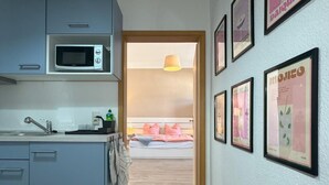A compact kitchen with a microwave, sink, and cabinets. A hallway leads to a bedroom with a bed and a nightstand. - Ferienwohnung Plohn_immo-franzi (Lengenfeld)