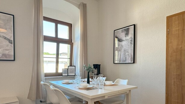 A dining area with a white table set for four, wooden chairs, and framed artwork on the walls. - Ferienwohnung Plohn_immo-franzi (Lengenfeld)