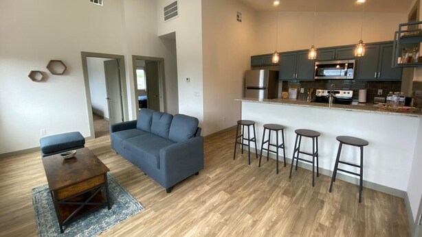 A modern living room with a grey sofa, a wooden coffee table, and a kitchen area with a bar stool set.
