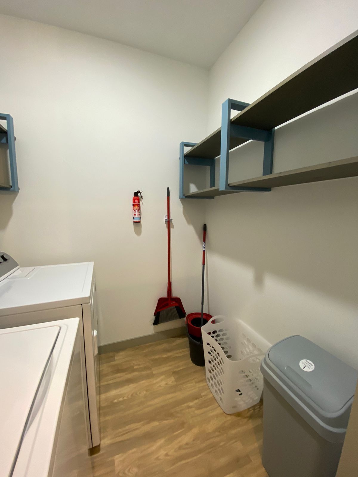 A laundry room with a washing machine, a broom, a dustpan, a trash bin, and a fire extinguisher.