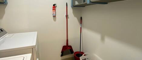 A laundry room with a washing machine, a broom, a dustpan, a trash bin, and a fire extinguisher.