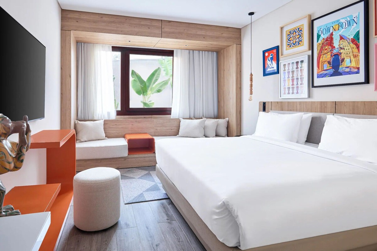 A hotel room with a large bed, a sofa, a desk, and a television. There are framed pictures on the wall and a window with curtains.