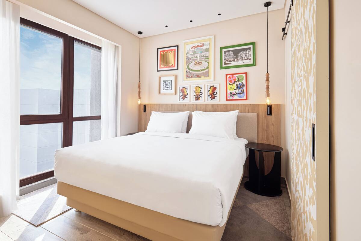 A modern hotel room with a large bed, a wooden headboard, and a large window. There are several framed pictures on the wall above the bed.