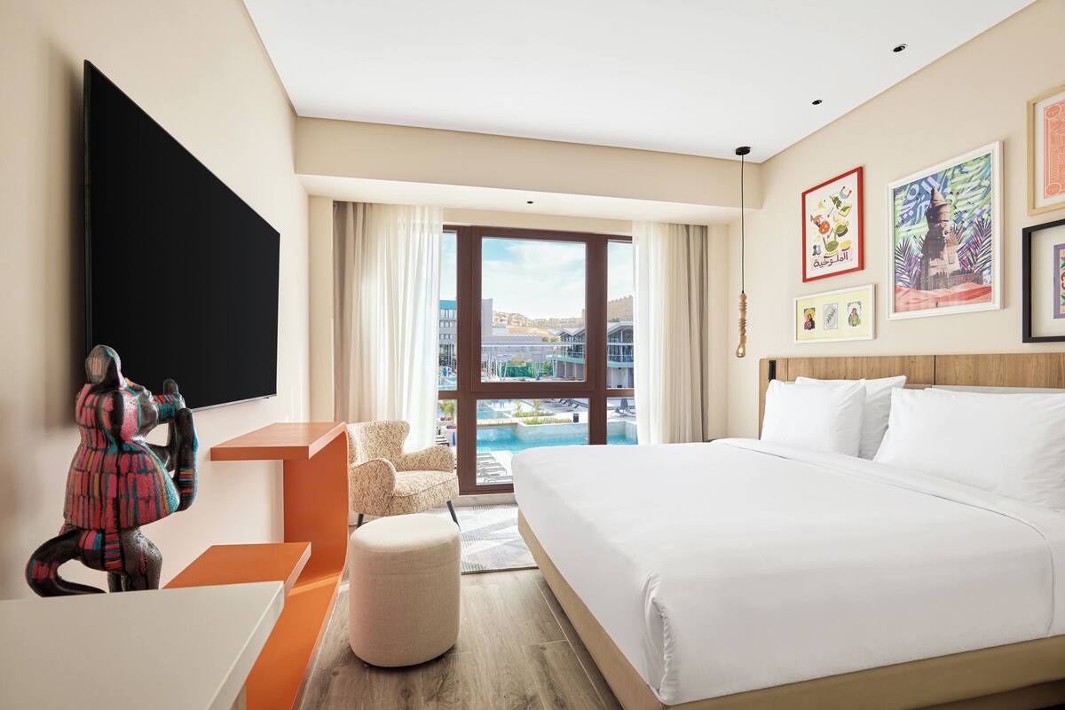 A hotel room with a large bed, a flat-screen TV mounted on the wall, a seating area with a chair and ottoman, and a view of a pool and buildings through the window.