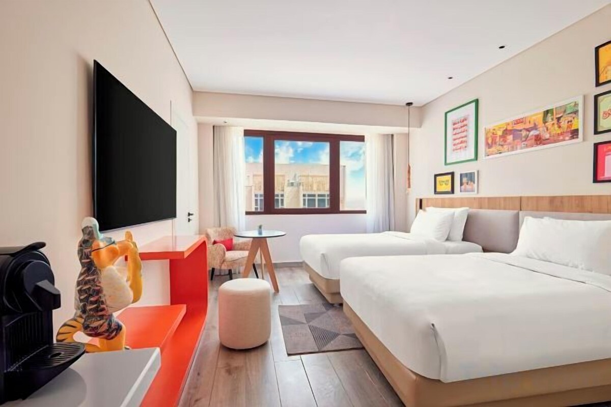 A hotel room with two beds, a flat-screen TV, a coffee machine, and a small table with a chair.