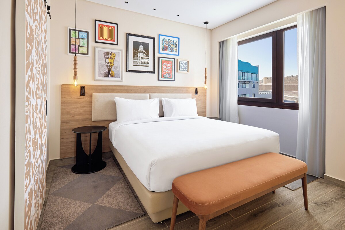 A hotel room with a large bed, a bench, a small table, and a view of the city through a window.