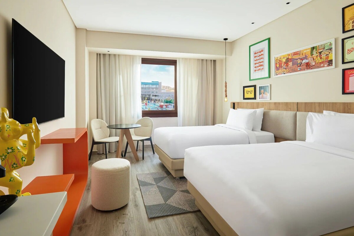 A hotel room with two beds, a TV, a desk with a coffee maker, a chair, and a window with a view of buildings.