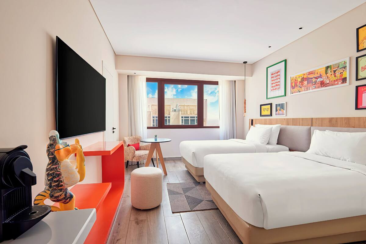 A hotel room with two beds, a TV, a coffee machine, a small table, and a window with a city view.