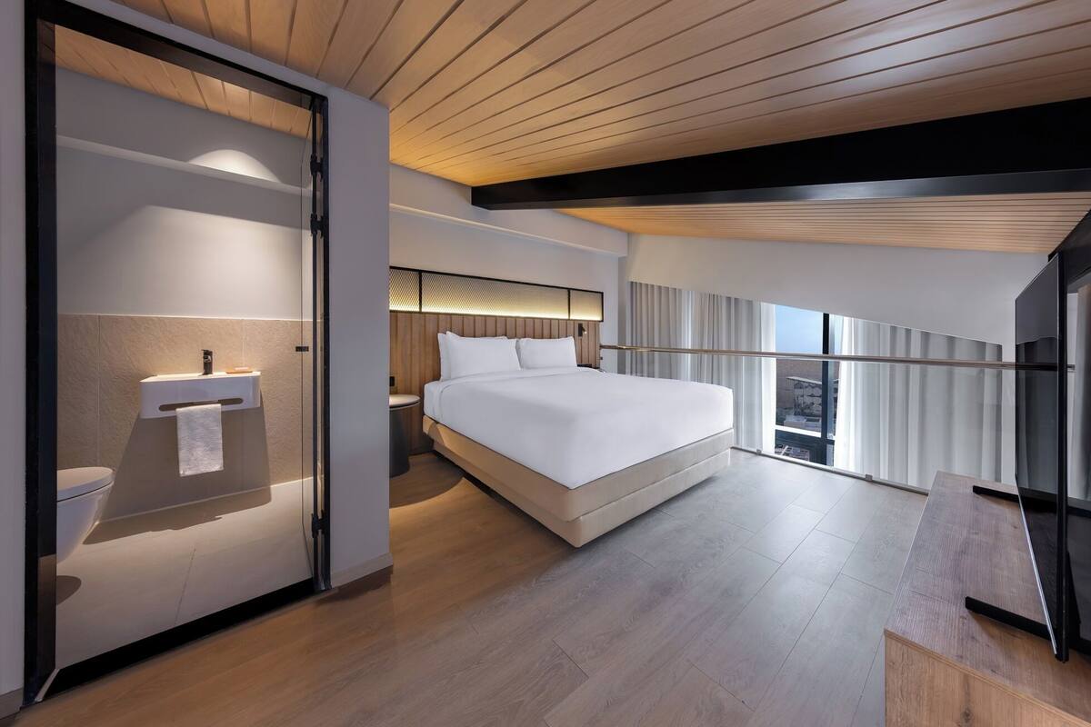 A modern hotel room with a large bed, a bedside table, a built-in TV, and a balcony with a view.