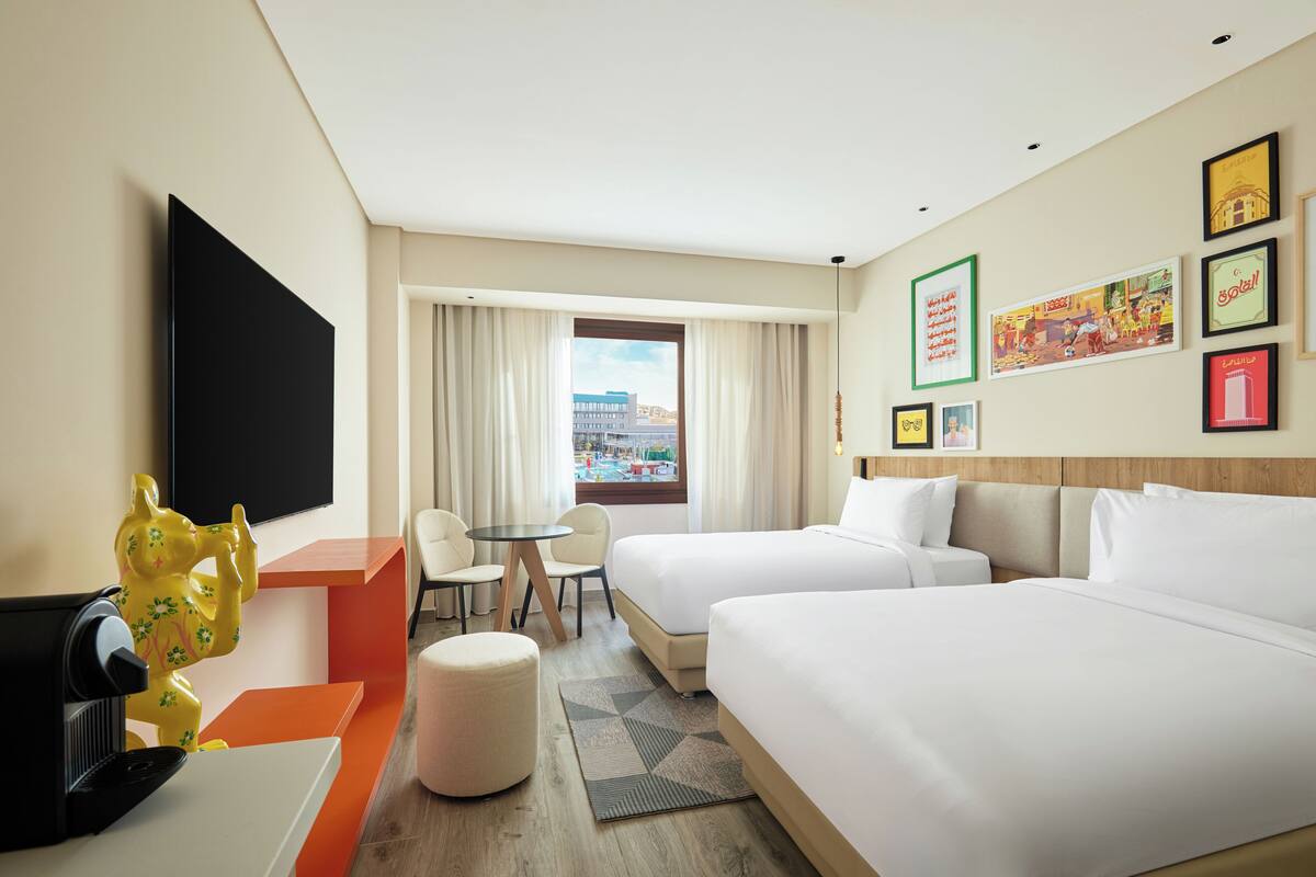 A hotel room with two beds, a TV, a desk, a chair, and a window with a view of buildings.