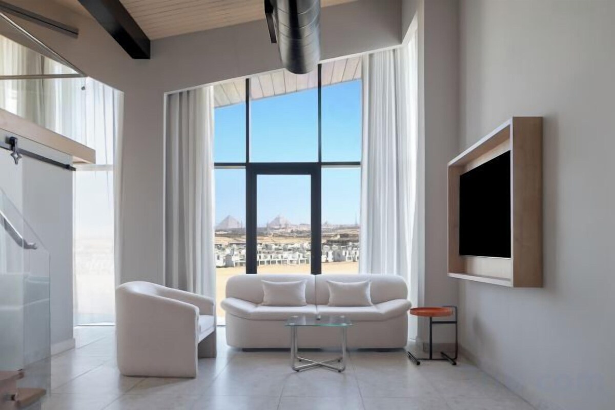 A modern living room with a large window, a sofa, a coffee table, and a TV mounted on the wall.