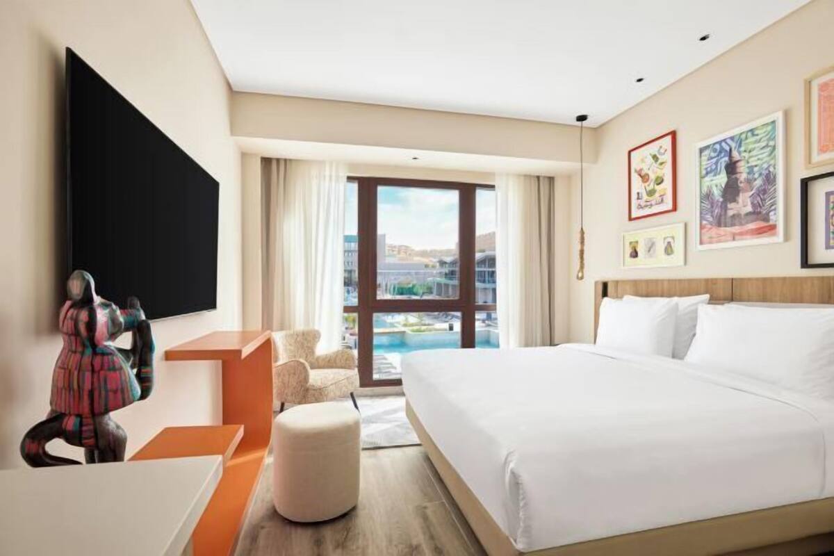 A hotel room with a large bed, a flat-screen TV, a seating area with a chair and ottoman, and artwork on the walls.