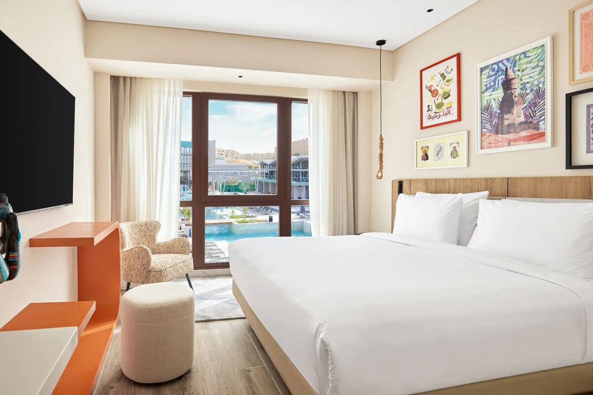 A hotel room with a large bed, a TV, a desk, a chair, and a view of a swimming pool and buildings.