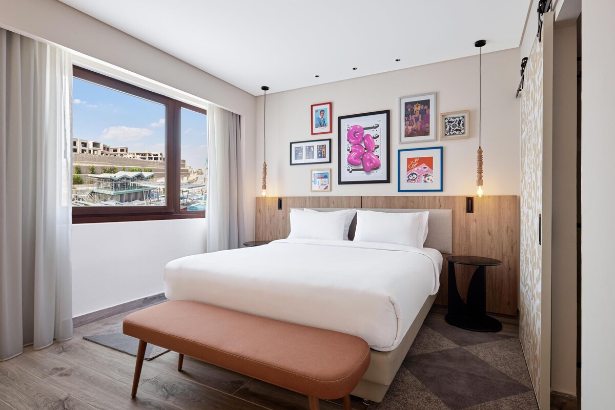 A modern hotel room with a large bed, a bench, a view of the city, and artwork on the walls.