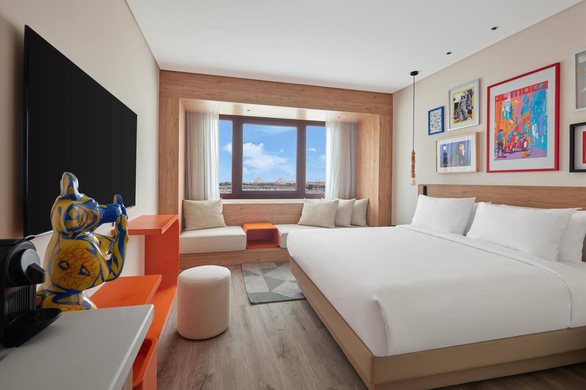A modern hotel room with a large bed, a seating area, a TV, and artwork on the walls.