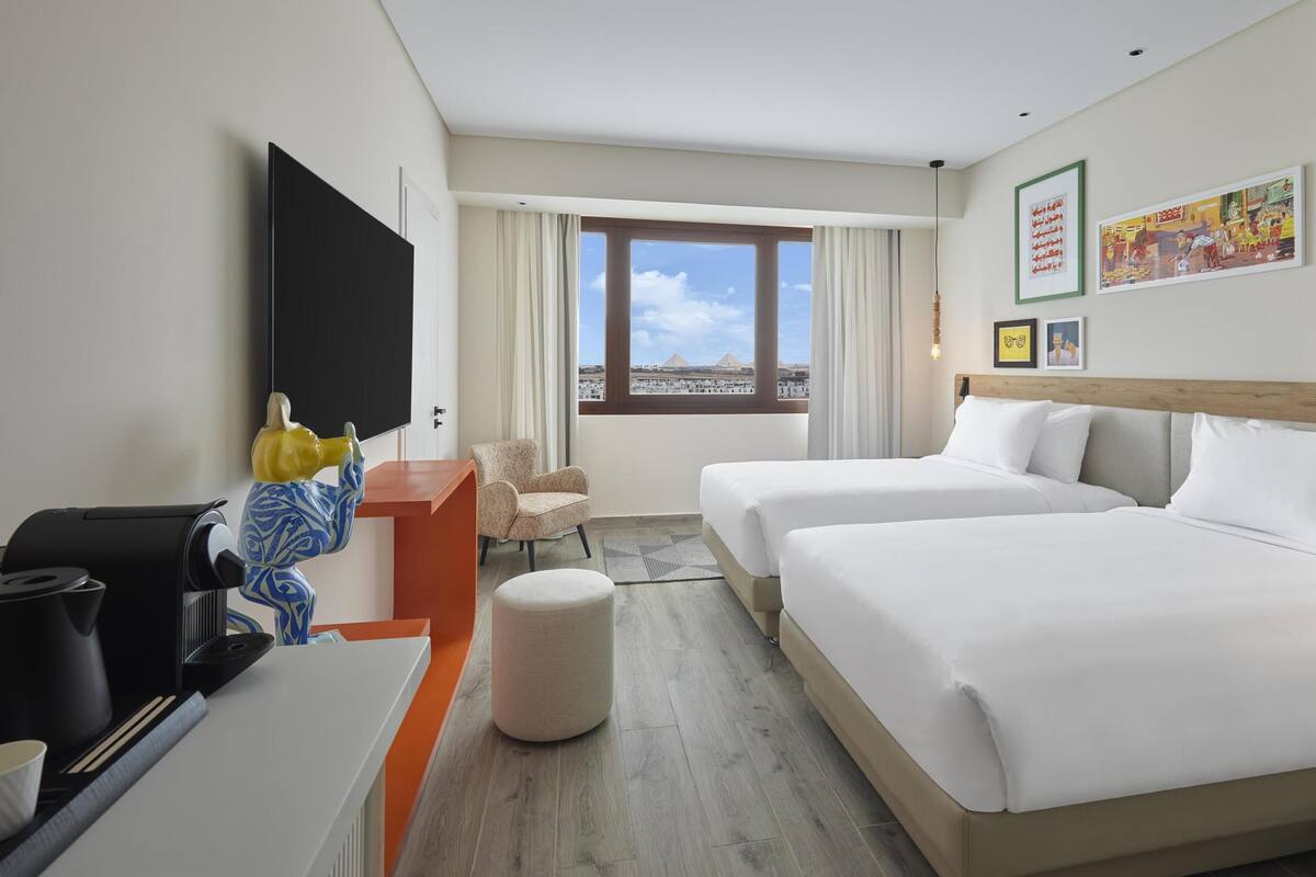 A hotel room with two beds, a desk with a coffee maker, a chair, and a large window with a view of a pyramid.