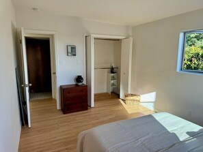 A bedroom with a bed, wooden flooring, a dresser, a closet, and a window with a view of greenery. - Mid-Century Retreat in the Heart of Encinitas (Encinitas)