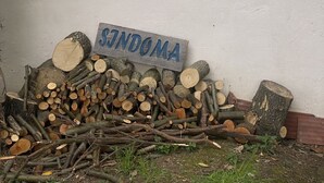 Une pile de bois de chauffage est appuyée contre un mur, à côté d’un carillon suspendu et d’une pancarte sur laquelle on peut lire « SINDOMA ».